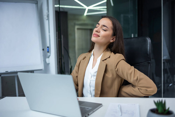 caucasian businesswoman working with laptop in cabinet feeling severe back pain while sitting on office chair indoors. - Foto, Bild