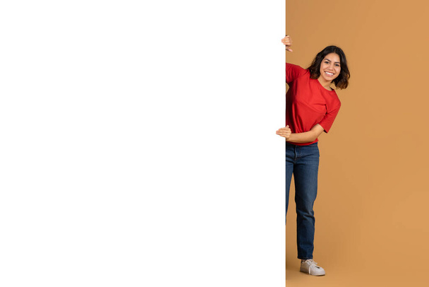 Joyeux jeune femme arabe posant à côté de l'espace énorme blanc blanc panneau publicitaire maquette copie, arrière-plan studio coloré. Offre ou offre intéressante, annonce - Photo, image