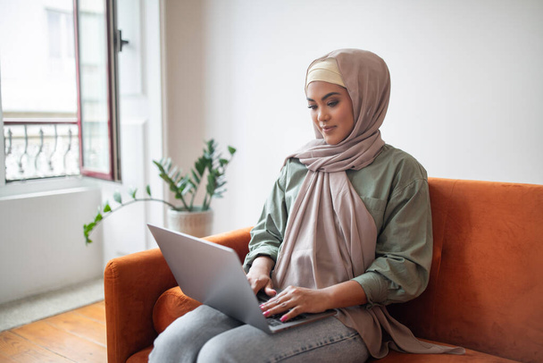 Trabajar y aprender. La mujer islámica milenaria en pañuelo para la cabeza trabaja en su computadora portátil, combinando la carrera remota y el aprendizaje electrónico, sentada en el sofá con computadora en la acogedora sala de estar moderna en casa - Foto, imagen