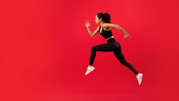 Ταιριάζει νεαρή γυναίκα σε μαύρο αθλητικό ντύσιμο πηδά δυναμικά σε έντονο κόκκινο φόντο, ενσωματώνοντας δύναμη και κίνηση. Πλευρική άποψη της αθλήτριας που τρέχει στον αέρα. Πανόραμα, αντίγραφο χώρου - Φωτογραφία, εικόνα