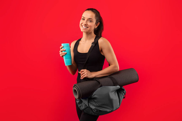 Happy sportovní tisíciletá žena drží fitness tašku s rohožkou a láhev vody, pózování v černém sportovním oblečení připraven k tréninku na červeném pozadí studia, s úsměvem na kameru - Fotografie, Obrázek