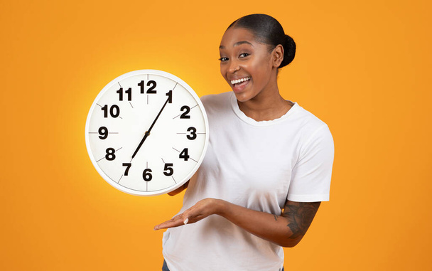 Usmívající se Afroameričanka drží velké hodiny, ukazuje dochvilnost a řízení termínu, pózuje nad oranžovým studiem na pozadí, usmívá se do kamery, ztělesňuje koncept času - Fotografie, Obrázek