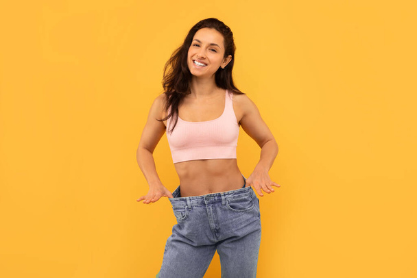 Result Of Successful Weight Loss. Cheerful Young Slim Lady Wearing Big Oversize Jeans To Compare Waist Size After Slimming, Posing Over Yellow Studio Backdrop, Smiling To Camera - Photo, Image