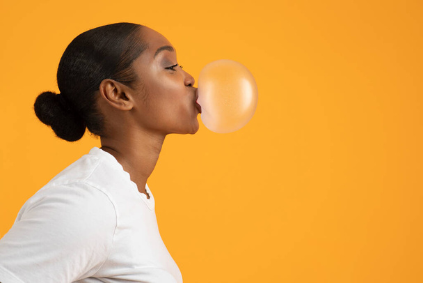 Profil de la dame afro-américaine souffle bubblegum pour le plaisir sur fond orange jaune vif. Restez insouciant concept. Bannière publicitaire avec espace libre pour le texte - Photo, image