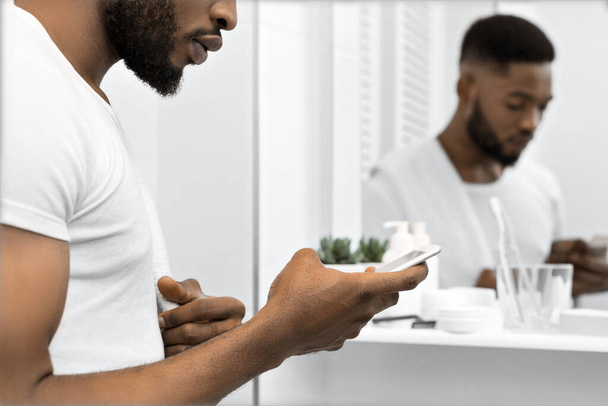 Knappe Afro man lezing bericht in smartphone in de badkamer. Man in badkamer concept - Foto, afbeelding