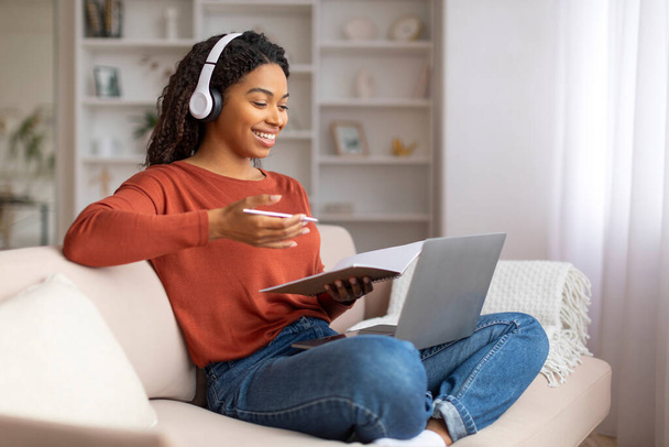 Sonriente joven mujer negra que usa auriculares multitarea en casa, escribiendo en un cuaderno mientras usa el ordenador portátil, mujer afroamericana feliz que asiste a un curso en línea o ve un seminario web, sentado en un sofá - Foto, imagen
