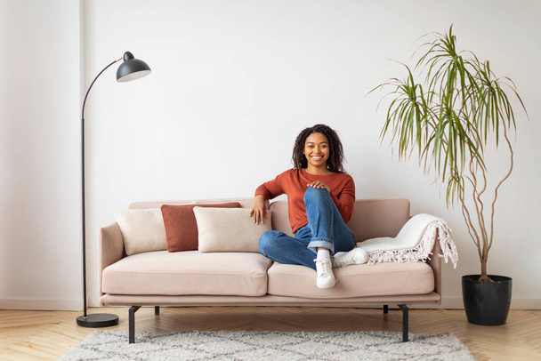 Nyugodt fiatal fekete nő örömteli mosollyal kényelmesen ül otthon a bézs kanapén, boldog ezeréves afro-amerikai hölgy pózol jól megvilágított nappaliban, laza eleganciát áraszt, másolja a helyet - Fotó, kép