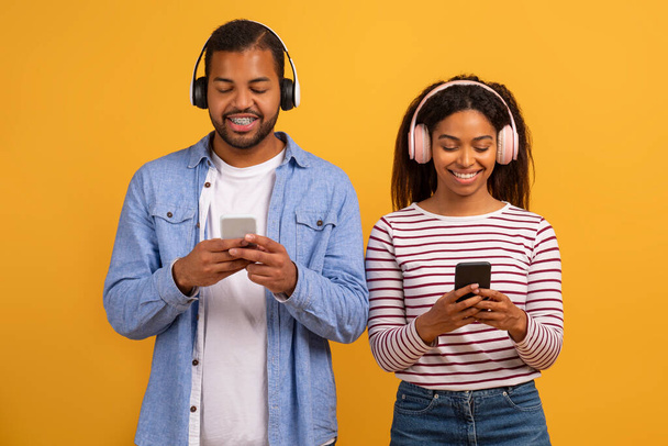Smiling Black Couple Wearing Wireless Headphones Using Modern Smartphones, Happy African American Man And Woman Listening To Music Online, Enjoying Favorite Song, Standing On Yellow Studio Background - Foto, immagini
