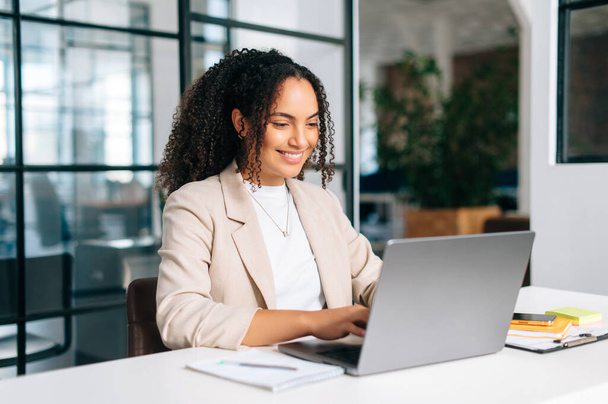 Positiv schöne lockige hispanische oder lateinamerikanische junge Frau in eleganter Kleidung, Unternehmerin, Büroangestellte, die an einem Laptop arbeitet, während sie an ihrem Arbeitsplatz im modernen Büro sitzt, lächelt - Foto, Bild