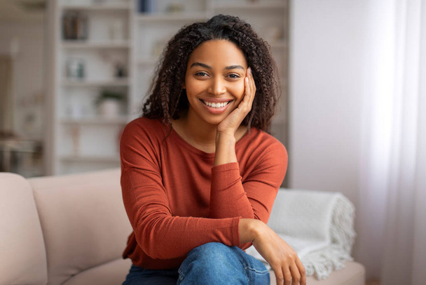 Retrato de una atractiva joven negra con una sonrisa cautivadora sentada casualmente en el sofá, mujer afroamericana feliz posando en una acogedora sala de estar en casa, reflejando un estilo de vida relajado y contenido - Foto, Imagen