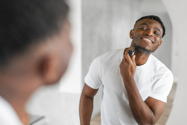 Happy knappe Afro-Amerikaanse man scheren met een elektrisch scheermes, geniet 's morgens routine in zijn moderne badkamer, benadrukt mannelijke huidverzorging en ontharing, zelfverzorging. Selectieve focus - Foto, afbeelding
