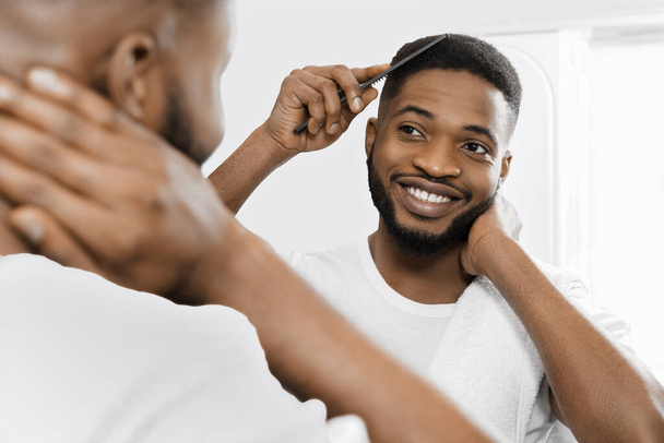 glimlachen afro-Amerikaanse man kammen haar en kijken in de spiegel in de badkamer. - Foto, afbeelding