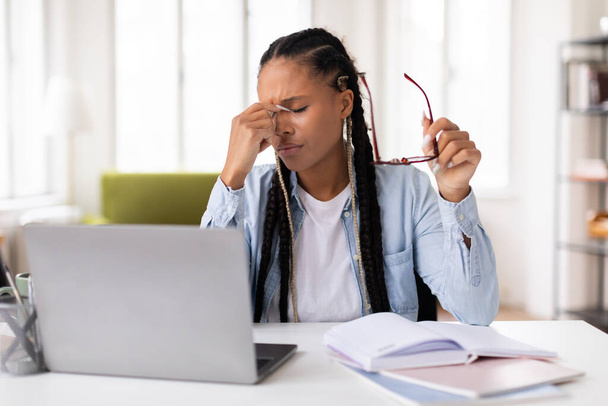 Сосредоточенная черная студентка чувствует стресс от головной боли, массирует носовой мост и держит очки во время учебы за ноутбуком на столе, домашний интерьер - Фото, изображение