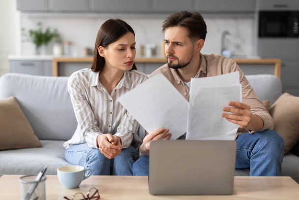 Worried young couple examines paperwork together, sitting on couch in front of open laptop and with cup of coffee on table, in living room home setting - Photo, Image