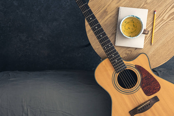 テーブルの上にアコースティックギター,メモ帳,紅茶のカップを備えたミュージカルバック. ギターを弾くこと,曲を書くことを学ぶことの概念. コピースペース. - 写真・画像