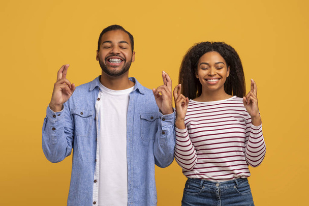 Veselý mladý černošky pár křížení prsty, dělat přání se zavřenýma očima, pozitivní afroameričan muž a žena žadonící o štěstí a štěstí, stojí spolu proti žlutému studiu na pozadí - Fotografie, Obrázek