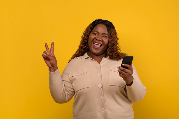 黄色い背景に隔離された平和なジェスチャーと笑顔を示す彼女の手でスマートフォンでベージュのシャツを着ているかなり長髪のアフリカ系アメリカ人女性. デートアプリ - 写真・画像