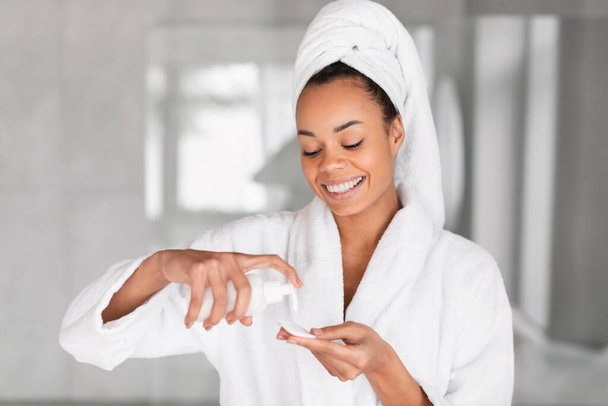 Fröhliche Afroamerikanerin mit eingewickeltem Handtuch auf dem Kopf, die Tonic auf Wattepad aufträgt und die Gesichtshaut mit strahlendem Lächeln pflegt, während sie im heimischen Badezimmer drinnen steht. Hautpflegeprodukte - Foto, Bild