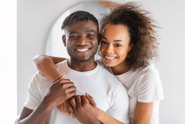 Retrato de feliz joven pareja negra vestida con camisetas blancas, abrazos en el ambiente del baño, sonriendo a la cámara, simbolizando el vínculo amoroso y momentos compartidos de autocuidado. Mimos familiares - Foto, imagen