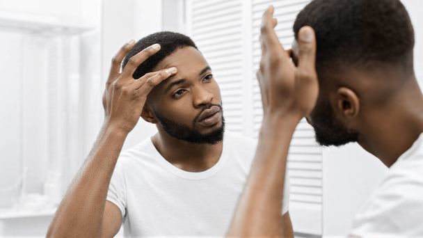Afro-Amerikaanse man raakt zijn gezicht, kijkt zichzelf in spiegel in de badkamer. Mans Skin problem concept - Foto, afbeelding