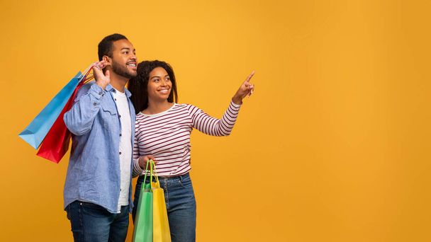 Nice Offer. Excited young black spouses with bright shopping bags pointing aside, indicating copy space, standing isolated over yellow background, african american couple enjoying seasonal discounts - Photo, Image