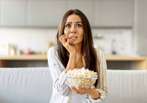 Contenuto d'urto. Paura giovane donna guardando la TV e mangiare popcorn a casa, terrorizzato Millennial Femmina mordere le unghie, emotivamente reagendo allo show televisivo, seduto sul divano in salotto - Foto, immagini
