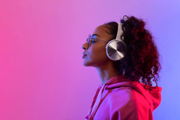 音楽で失われたワイヤレスヘッドフォンを持つ平和な黒人女性,ネオンピンクとブルーグラデーションの背景,サイドビュー,無料スペースを着用した女性 - 写真・画像