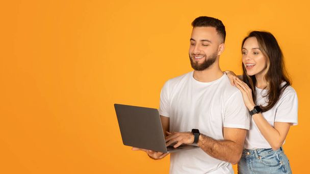 Jeune homme souriant tenant un ordinateur portable avec une femme attentive à ses côtés, tous deux portant des t-shirts blancs, apparemment engagés dans une découverte en ligne intéressante, sur un fond orange vif - Photo, image