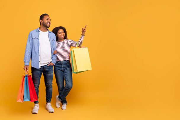 Seasonal Sales. Smiling Black Spouses Walking With Shopping Bags And Pointing Aside At Copy Space On Yellow Background In Studio, Happy Middle Eastern Couple Enjoying Shopper Deals, Panorama - Photo, Image