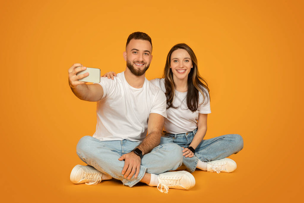 Encantado casal caucasiano sentado de pernas cruzadas no chão, tirando uma selfie com um smartphone, ambos vestidos com camisetas brancas e jeans azuis contra um fundo laranja alegre - Foto, Imagem