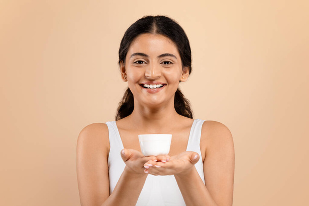 Meilleurs cosmétiques. Belle jeune femme indienne tenant pot avec crème hydratante et souriant à la caméra, attrayant Millennial femelle en blanc Top montrant un produit de beauté pour la peau, Copier l'espace - Photo, image