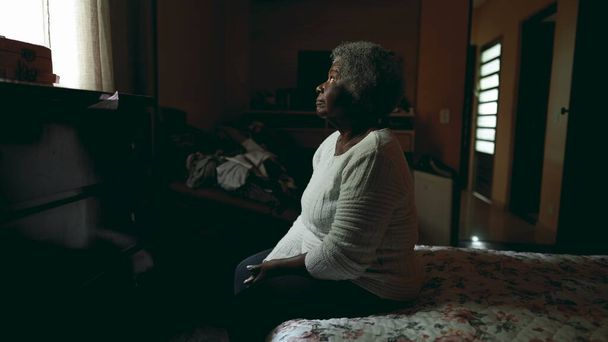 Μοναχική Νοτιοαμερικανίδα ηλικιωμένη κυρία που κάθεται δίπλα στο κρεβάτι με γαλήνιο βλέμμα στο παράθυρο μέσα στο αμυδρά φωτισμένο ταπεινό υπνοδωμάτιο που ατενίζει τη ζωή και τις αναμνήσεις του παρελθόντος. ηλικιωμένος 80 ετών - Φωτογραφία, εικόνα