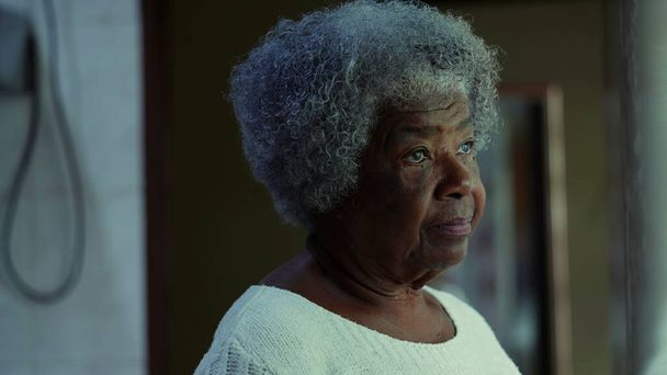 Μια στοχαστική ηλικιωμένη μαύρη γυναίκα της δεκαετίας του 80 στέκεται στο σπίτι με το χέρι στο πηγούνι μελετώντας βαθιά για τα προβλήματα, σκεπτική έκφραση της Αφρο-Αμερικανίδας γκρίζας κομμώτριας - Φωτογραφία, εικόνα