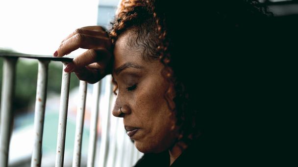 Jedna przygnębiona czarna kobieta smutek i rozpacz z zamkniętymi oczami opierając się na metalowym barze na balkonie, profil zbliżeniowy twarz zdesperowanej osoby w średnim wieku smutek - Zdjęcie, obraz