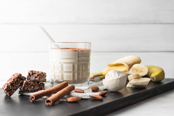 タンパク質ドリンク付きのガラス,バナナ,タンパク質バー,アーモンドナッツ,シナモンスティックを測定するタンパク質粉末の木製ボード上の健康的なミルクシェークスムージー. - 写真・画像