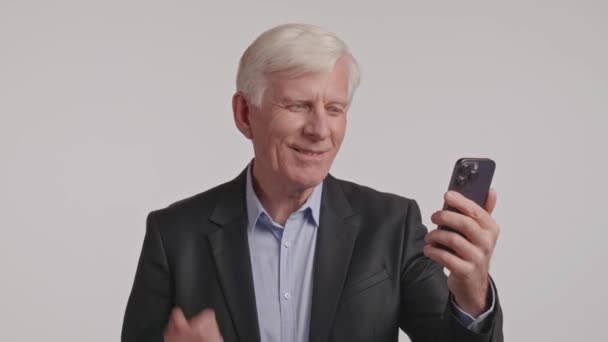 Een oudere man in formele kleding met een blazer glimlacht terwijl hij een mobiele telefoon in zijn hand houdt. Zijn duim is op het scherm, toont een gebaar van het gebruik van het communicatieapparaat - Video