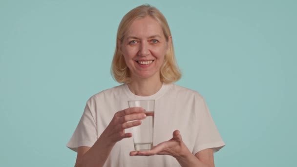 Una mujer sostiene un vaso de agua, sonríe con la boca y muestra un gesto feliz. Su mandíbula está relajada mientras su manga se cae, y su dedo y pulgar están posicionados de una manera divertida - Imágenes, Vídeo