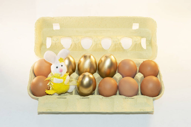 おもちゃのウサギ,イースターバニーが付いている茶色および金の卵が付いている段ボール紙のペーパー皿. イースターのための装飾的な染められた卵が付いているパック. 卵の値上げについて。 卵箱。 休日のための準備. - 写真・画像