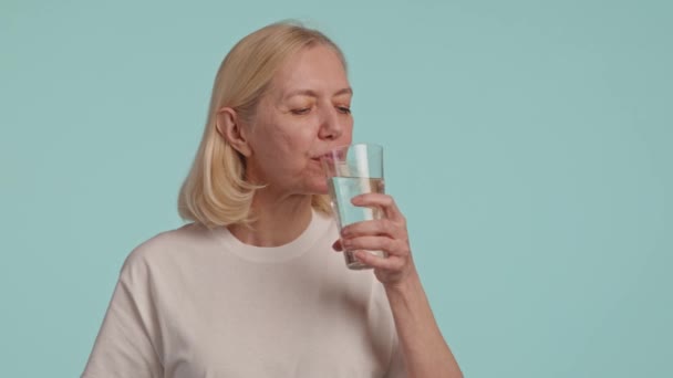 Una donna beve felicemente un bicchiere d'acqua, con un sorriso sul viso e il braccio alzato. Il dito toccando delicatamente il bordo del vetro, ciglia svolazzanti, su uno sfondo blu calmante - Filmati, video