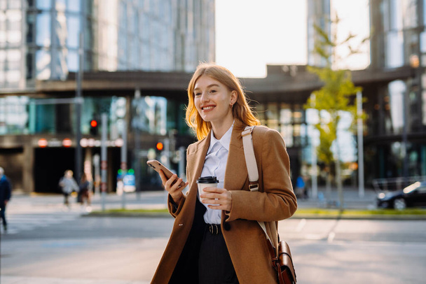 Pewna siebie bizneswoman spacerująca z telefonem i kawą wzdłuż centrum miasta, pozująca na ulicy, w drodze do biura - Zdjęcie, obraz