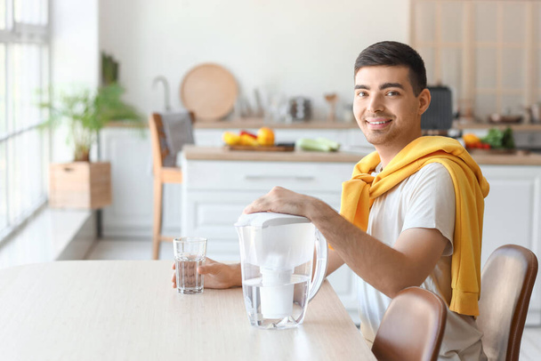 Młody człowiek z dzbankiem filtracyjnym i szklanką świeżej wody siedzi przy stole w kuchni - Zdjęcie, obraz
