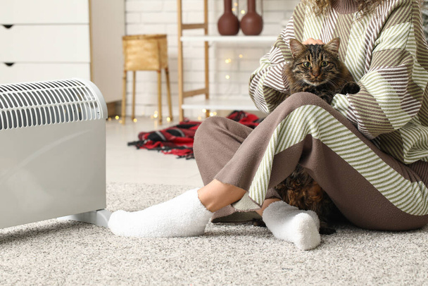 Žena s roztomilou kočkou sedí na podlaze v blízkosti elektrického ohřívače doma - Fotografie, Obrázek