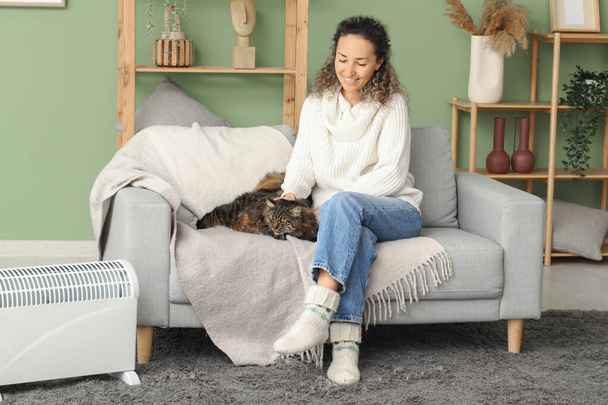 Happy mature woman with cute cat sitting on sofa near electric heater at home - Photo, Image
