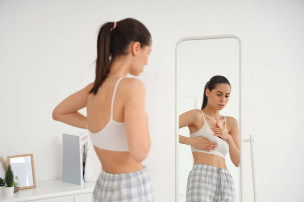 Νεαρή γυναίκα τσεκάρει το στήθος της κοντά στον καθρέφτη στην κρεβατοκάμαρα. Έννοια ευαισθητοποίησης για τον καρκίνο - Φωτογραφία, εικόνα