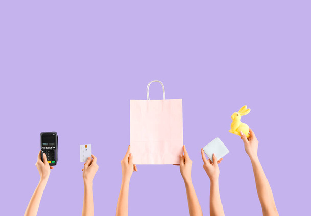 Kobiety z terminalem płatniczym, torbą na zakupy, kartą kredytową i portfelem na liliowym tle. Sprzedaż wielkanocna - Zdjęcie, obraz