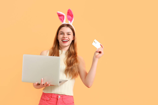 Jonge vrouw in konijnenoren met laptop en creditcard op oranje achtergrond. Pasen Verkoop - Foto, afbeelding