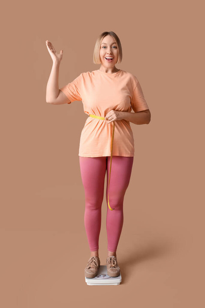Glückliche junge Frau mit Maßband um die Taille, die auf einer Waage vor beigem Hintergrund steht. Konzept zur Gewichtsreduktion - Foto, Bild