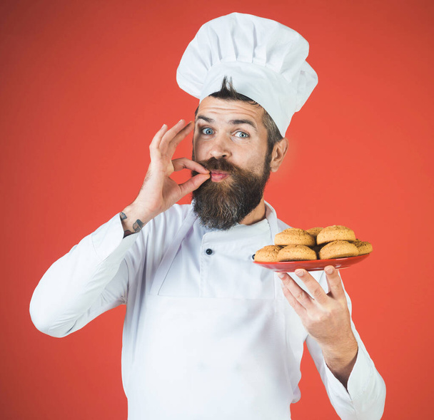 Αρσενικό σεφ με πιάτο μπισκότα βρώμης δείχνει ok υπογράψει. Χαμογελαστή σεφ, μάγειρας ή φούρναρης με χειρονομία έγκρισης γεύσης. Γενειοφόρος άνδρας με καπέλο σεφ και στολή με σπιτικά μπισκότα βρώμης δείχνει νόστιμο σημάδι - Φωτογραφία, εικόνα