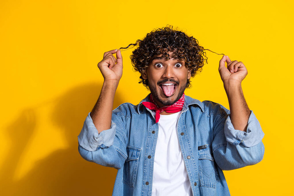 Photo of attractive young man hold curly strands tongue out dressed stylish denim clothes red scarf isolated on yellow color background. - Zdjęcie, obraz
