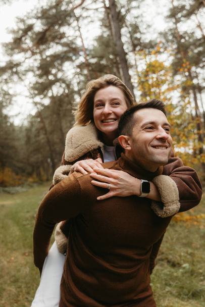 Erwachsenes Paar vergnügt sich im Herbstpark im Freien, glücklicher Mann trägt Frau auf dem Rücken in den Wald - Foto, Bild